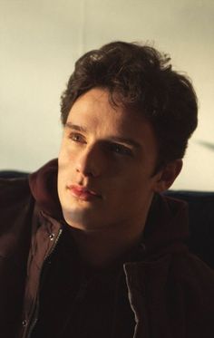 a young man with curly hair wearing a brown jacket and looking off to the side