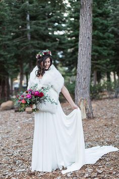 a woman standing in the woods wearing a white dress and fur stole with flowers on her head