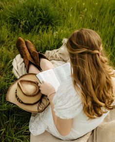 a woman sitting in the grass reading a book and holding a hat on her lap