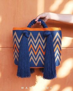 a hand holding a blue and orange bag with tassels on the handles, hanging from a wall