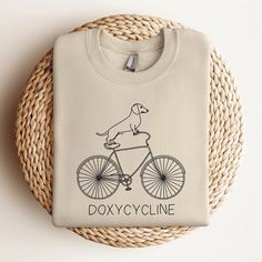 Doxycycline Pharmacy Pun Sweatshirt | Dachshund on Bicycle | Pharmacists Shirt | Veterinary | Pharmacy Gift | Gift for Pharmacists & Technicians PRODUCT DESCRIPTION: Ideal for any situation, a unisex heavy blend crewneck sweatshirt is pure comfort. These garments are made from polyester and cotton. This combination helps designs come out looking fresh and beautiful. The collar is ribbed knit, so it retains its shape even after washing. There are no itchy side seams on these sweaters.  - 50% cott Funny Shirt Women, Pharmacist Outfit, Veterinary Pharmacy, Pharmacy Shirt Ideas, Pharmacy Technician Shirts, Pharmacy T Shirt Ideas, Vet Sweatshirts, Funny Nurse Sweaters, Pharmacist Shirt