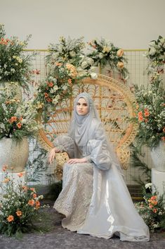 a woman wearing a hijab sitting on a wicker chair in front of flowers