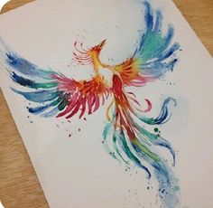 a colorful bird painted on white paper sitting on top of a wooden table next to a pen