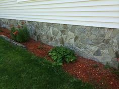 a stone wall next to a house with red flowers in the foreground and green grass on the other side