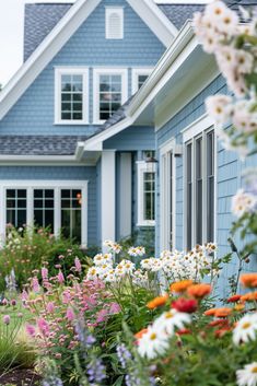 a blue house with white windows and lots of flowers in the foreground, on a sunny day