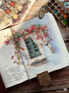 an open book sitting on top of a wooden table next to watercolor paints and brushes