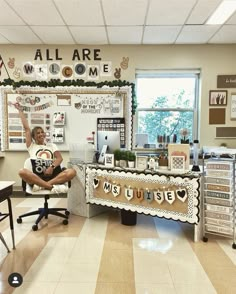 a woman sitting in a chair with her arms up and legs crossed, next to a bulletin board that says all are welcome