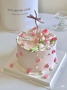 a white cake with pink flowers and a dragonfly on top is sitting next to a new mood cake box