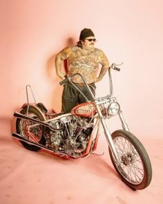 a man standing next to a motorcycle with tattoos on his arm and leg, in front of a pink wall