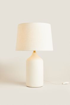 a white table lamp with a beige shade on the base and a cord plugged in