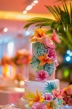 a wedding cake decorated with tropical flowers and greenery on a table in a banquet hall