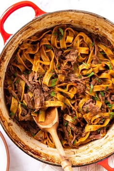 a pot filled with beef and noodles on top of a white table cloth next to a wooden spoon