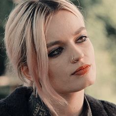 a woman with blonde hair and piercings looks off into the distance while wearing a black jacket