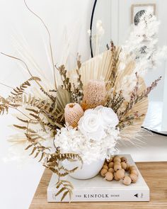 a white vase with flowers on top of a wooden table next to a book and mirror