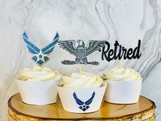 three cupcakes with frosting and an air force cake topper on a wooden stand