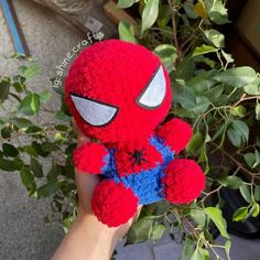 a hand holding a red and blue spiderman stuffed animal in front of green leaves