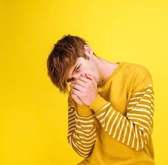 a young man covers his face with his hands while sitting on a stool against a yellow background