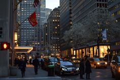 people are walking on the sidewalk in front of tall buildings at night with cars driving down the street