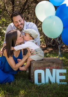 a man and woman kiss their son as they sit on a crate with balloons in the background