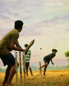 three young men playing cricket on a field at sunset or dawn with the sky in the background