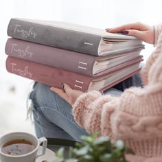 a woman is holding three books in her hands while sitting next to a cup of tea