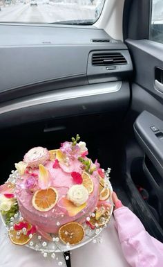 a person holding a cake in the back seat of a car with flowers and fruit on it