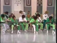 several children are sitting at desks in green uniforms and wearing white shirts, with their hands on their hips