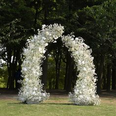 an arch made out of white flowers in the middle of a field with trees behind it