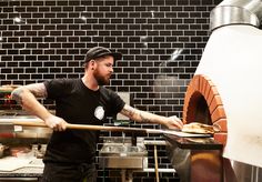 a man in black shirt holding a pizza on top of a wooden spatula next to an oven