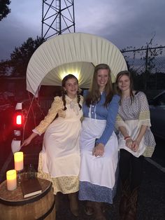 three women dressed in period clothing pose for a photo under an umbrella with candles on it