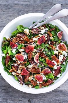 a white bowl filled with salad topped with figs and nuts next to two silver serving utensils