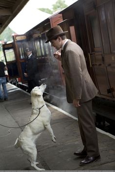 a man standing next to a train with his dog on a leash looking up at him