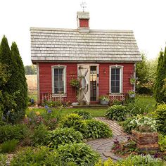 a small red house surrounded by greenery and flowers in the front yard, with a brick walkway leading to it