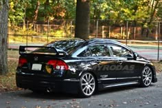 a black car is parked in front of a fence and trees with leaves on the ground