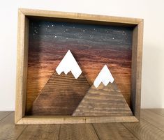 two mountains are cut out of wood in a shadow box on a wooden table with a white wall behind them