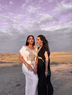 two beautiful women standing next to each other on a dirt field with clouds in the background