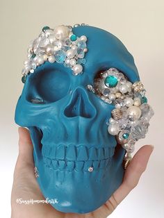 a hand holding a blue skull with pearls and jewels on it's head in front of a white background