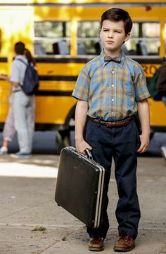 a young boy holding a suitcase in front of a school bus