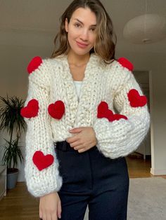 a woman wearing a white sweater with red hearts on the sleeves and cuffs, standing in front of a living room