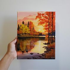 a hand holding up a painting of a sunset over a lake with trees in the background