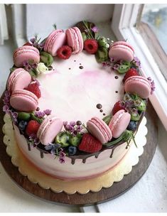 a cake decorated with macaroons and strawberries on a wooden platter next to a window