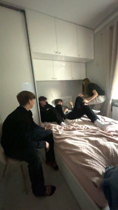 three people sitting on a bed in a room with white walls and flooring, one person is leaning over the edge of the bed