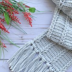 a crocheted scarf with red flowers and greenery next to it on a white wooden surface