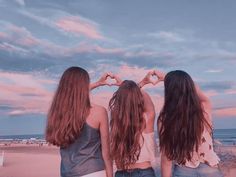 three young women standing on the beach making heart shapes with their hands while looking at the ocean