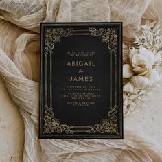 an elegant black and gold wedding card with white flowers on the table next to it