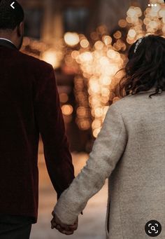 a man and woman holding hands while walking down the street with lights in the background