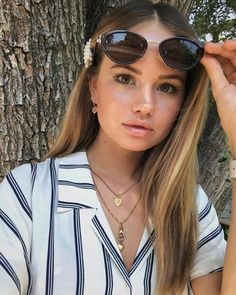 a woman wearing sunglasses standing in front of a tree with her hand on her head