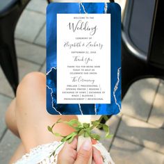 a person holding up a blue and white wedding card with greenery in it's hand