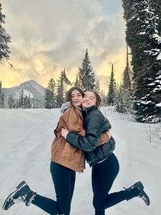 two women hugging each other in the snow with trees and mountains in the back ground