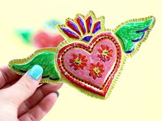 a hand holding up a heart shaped brooch with wings and flowers painted on it